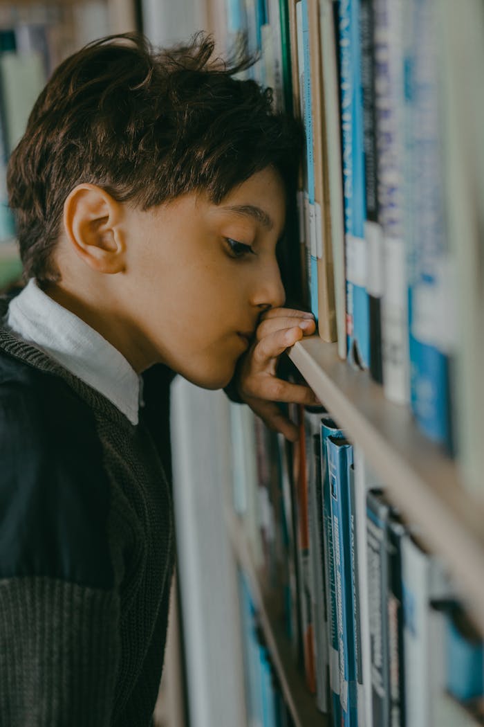 A young boy in a library, appearing thoughtful and reflective, leaning against bookshelves.
